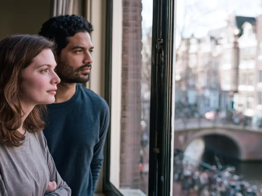 Couple standing close together at a window, one partner turned slightly away from the other